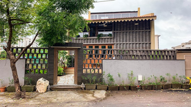 Street facade with perforated brick screen walls, planted bed, and tree beside the entry gate under overcast sky
