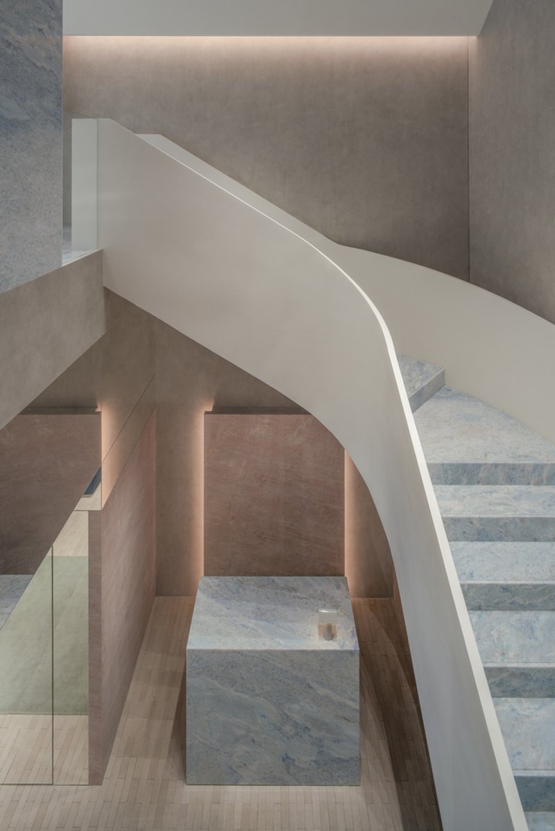 Curved white staircase with marble treads rising between pink stone walls under recessed linear lighting