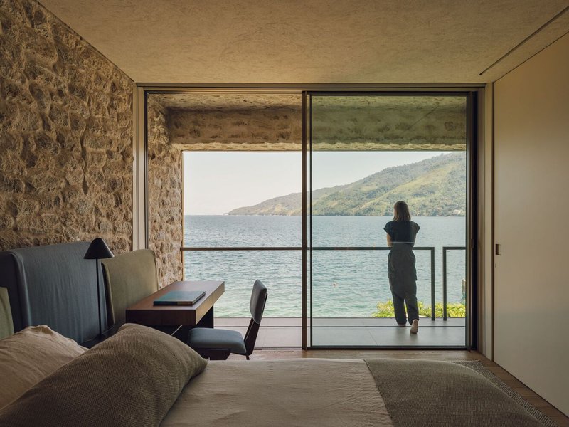 Bedroom with rough stone walls and floor-to-ceiling glazing framing a figure on the balcony overlooking the lake