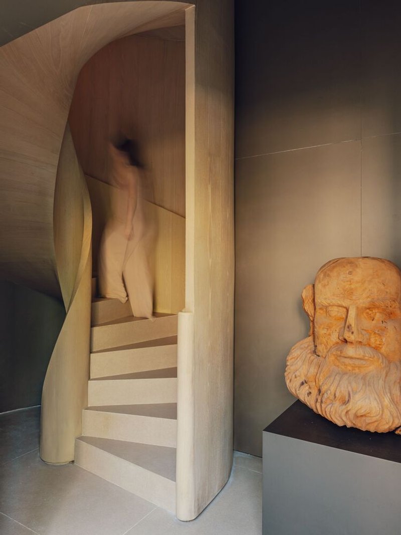 Curved timber staircase with sculptural handrail beside a terracotta bust on a plinth