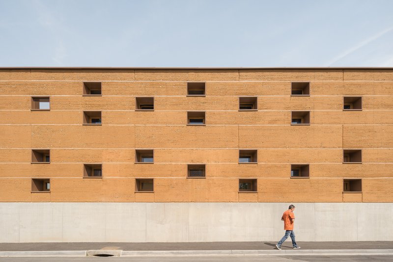 Street facade: long rammed earth wall with scattered small square windows, figure walking past