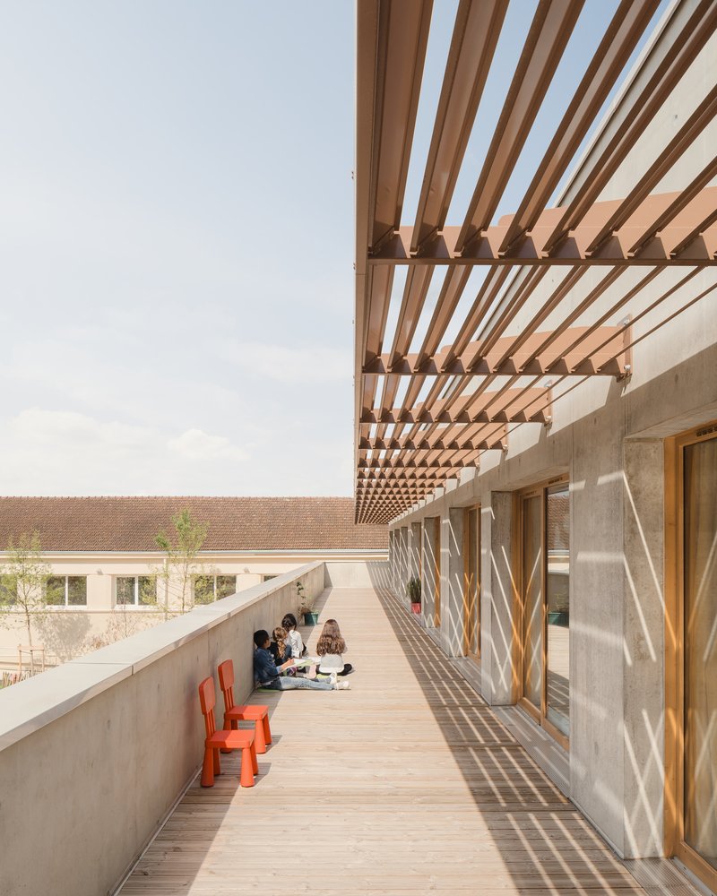 Upper walkway: timber louvre pergola providing shade, concrete parapet, students sitting, red chairs, rammed earth wall behind