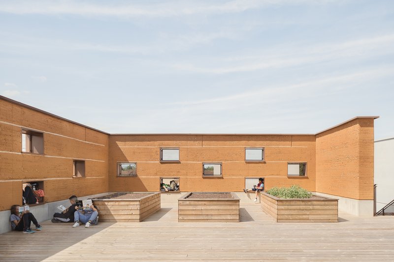 Rooftop courtyard: rammed earth walls with scattered windows, timber bench planters, students sitting and talking