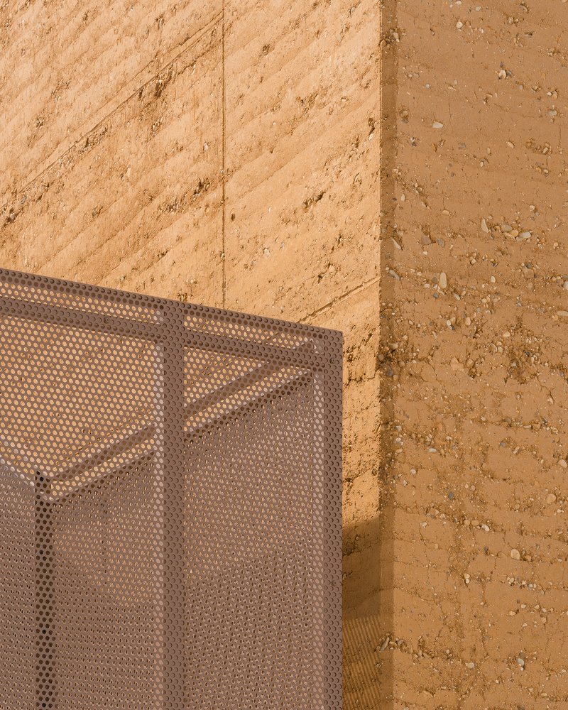 Detail: rammed earth wall corner meeting perforated metal mesh screen, warm afternoon sunlight on the earth texture