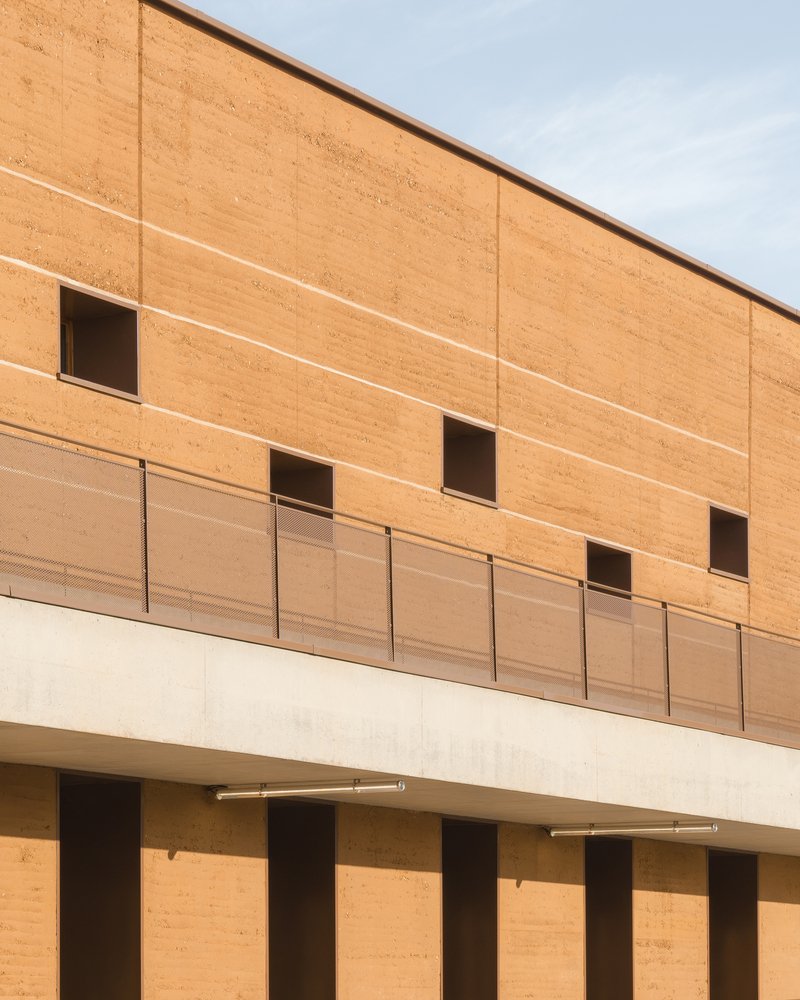 Facade: rammed earth upper floor with metal panel balustrade, concrete pilotis at ground level creating covered play area