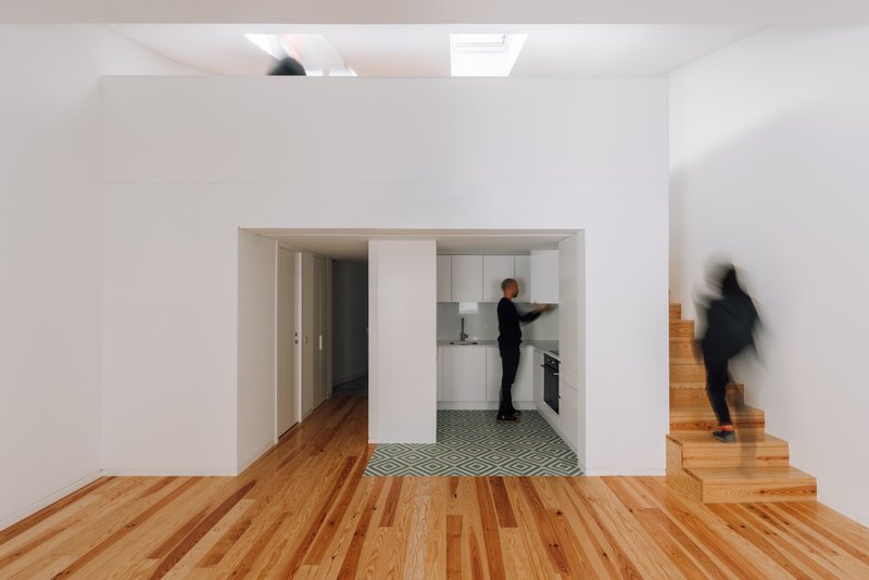 White interior room with timber floors looking through framed openings to a kitchen as a person ascends the stair