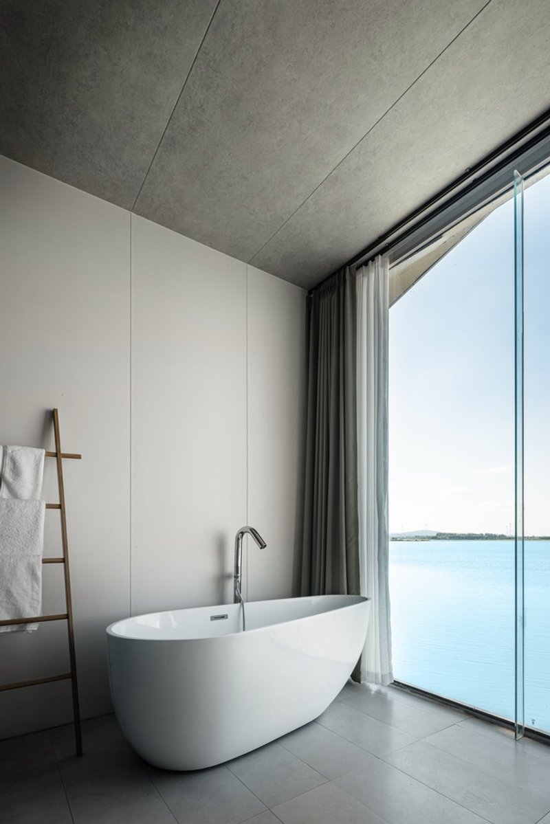 Freestanding bathtub positioned beside floor-to-ceiling glazing overlooking a calm blue lake