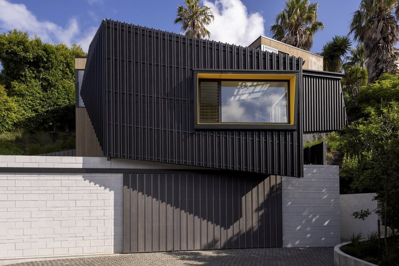 Vertical corrugated metal facade with yellow-framed window above white block retaining wall and palm trees