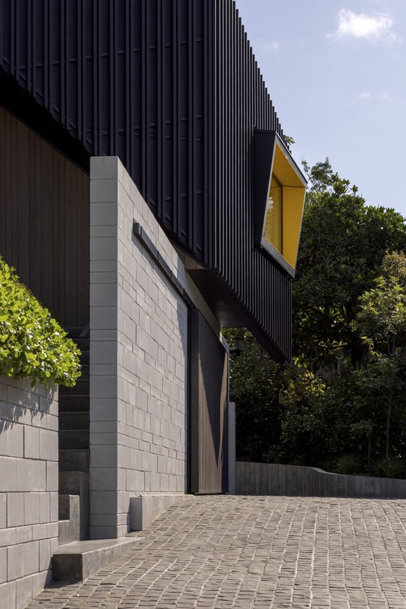 Entry courtyard showing corrugated metal volumes, yellow window frame, and concrete block walls meeting cobbled paving