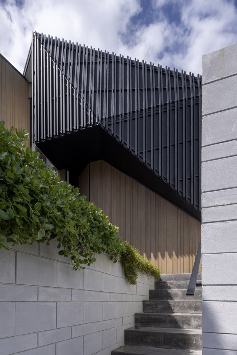 Cantilevered black vertical screen box above a concrete retaining wall with planted greenery