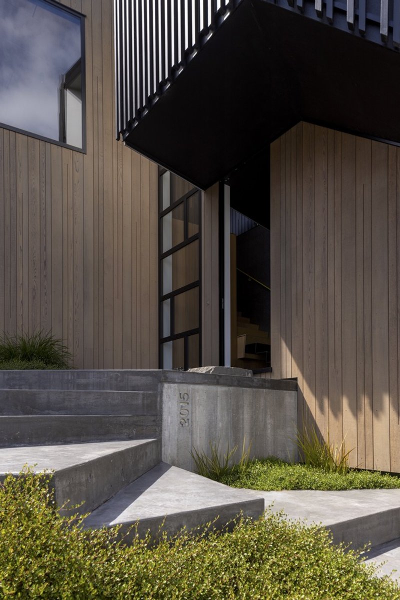 Entry sequence with concrete steps and black steel framed doorway beneath a cantilevered upper volume