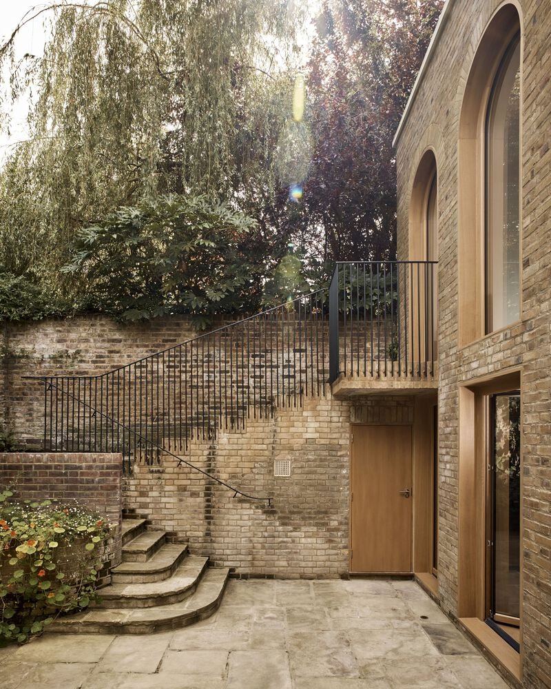 Courtyard with brick stairs climbing boundary wall beneath overhanging willow and copper beech trees