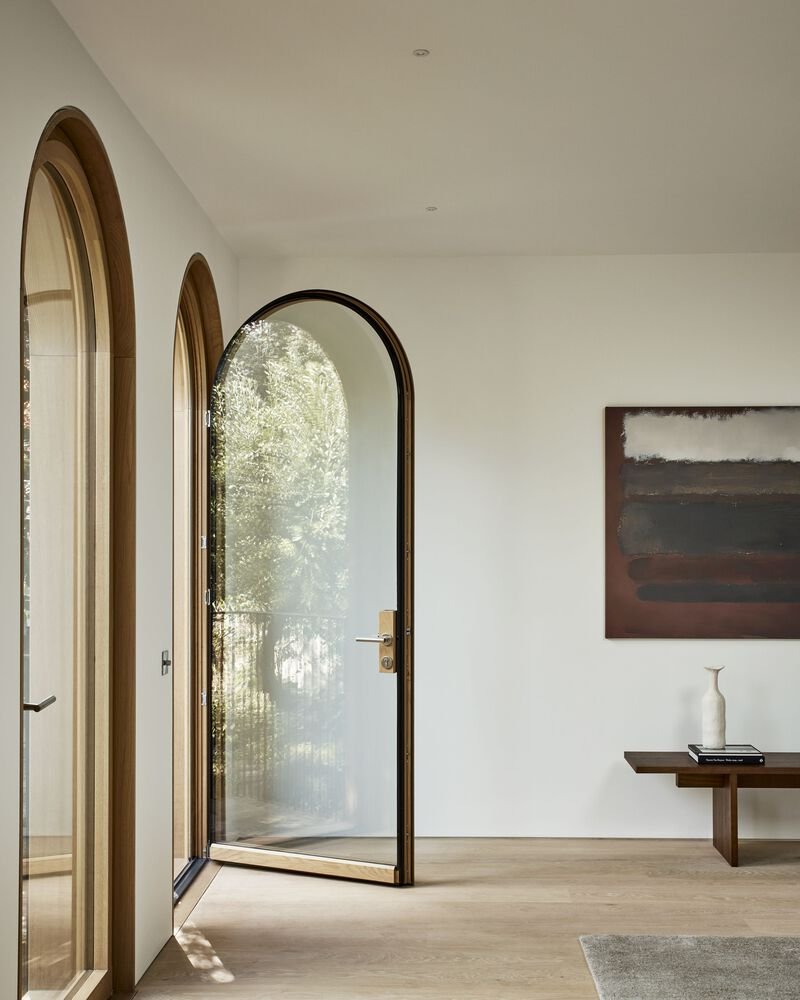 Open arched glass door with timber and bronze frame revealing sunlit garden foliage beyond
