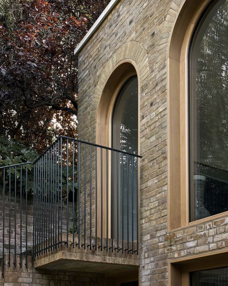 Yellow brick facade with tall arched window and metal-railed balcony beneath a dark tree canopy
