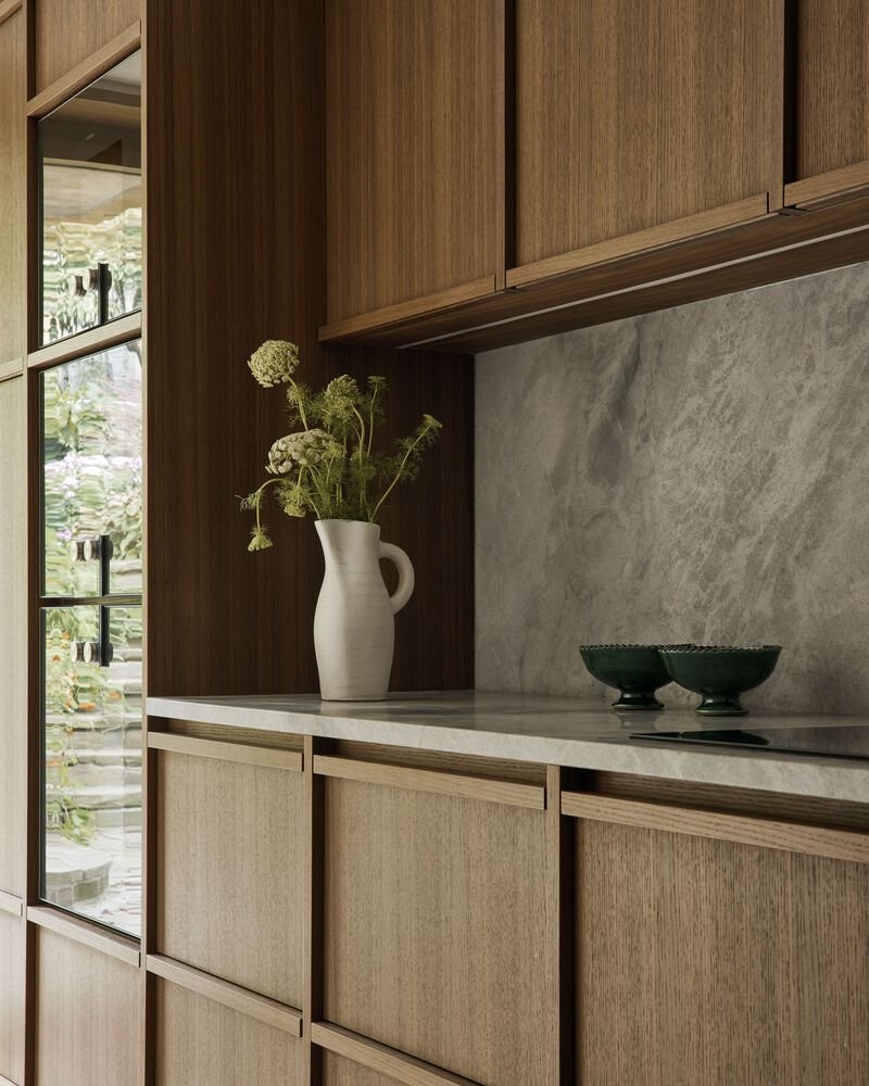 Kitchen counter with timber cabinetry, grey stone worktop and backsplash, overlooking a courtyard garden