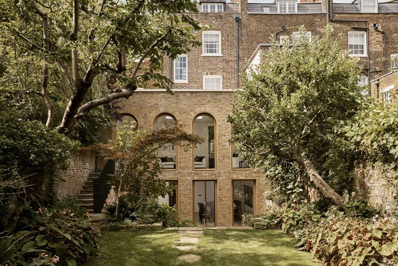 Garden facade with arched window openings in buff brick, framed by mature trees and lawn