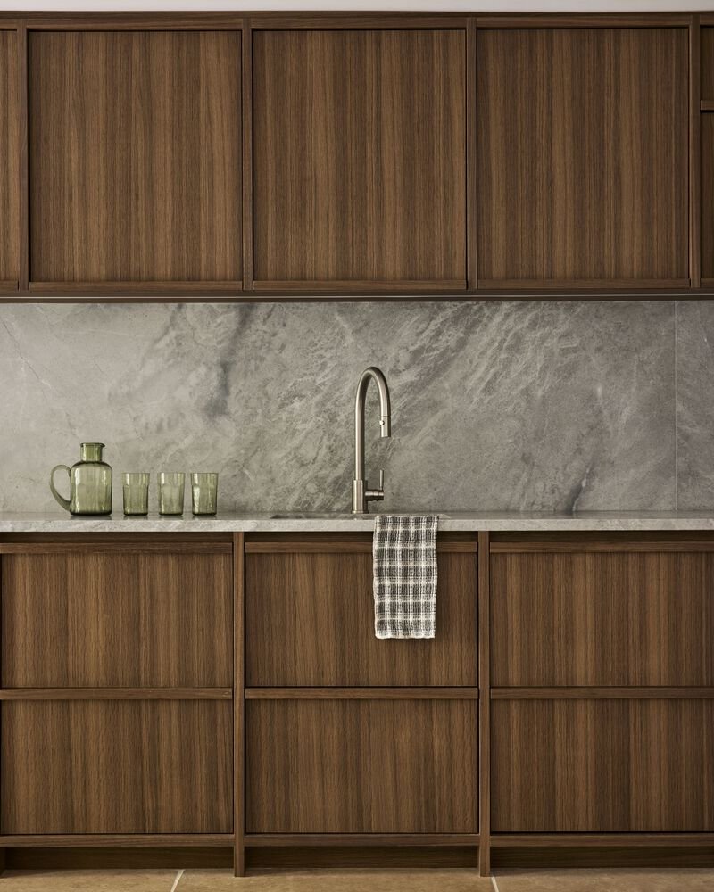 Kitchen sink detail with grey veined marble backsplash below walnut veneer cabinetry and glassware