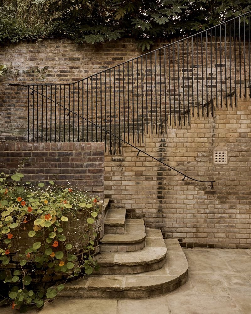 Curved stone staircase with metal balustrade against a buff brick wall, bordered by planted beds