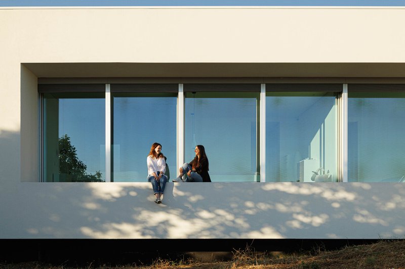 Cantilevered terrace with two seated figures behind floor-to-ceiling glass under dappled tree shadows