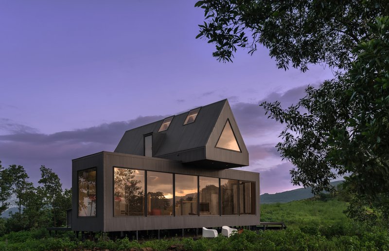 Gabled volume with glazed ground floor and dark pitched roof framed by trees at dusk