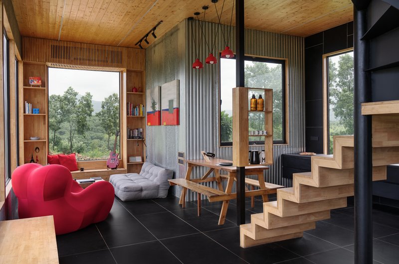 Open-plan living space with cantilevered timber stair, corrugated metal wall, and wooden ceiling under overcast sky
