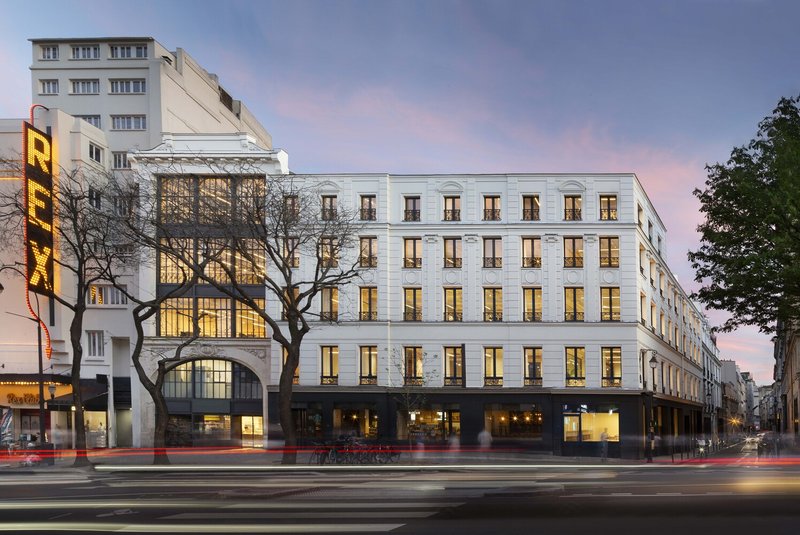 Street facade at dusk: restored Haussmann building with illuminated ground-floor steel and glass windows, Grand Rex cinema next door