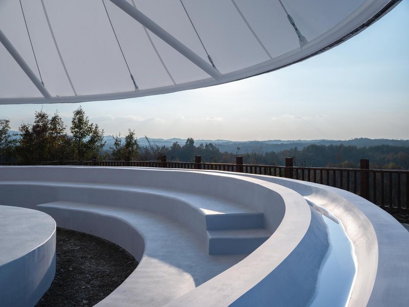 Curved concrete amphitheatre seating beneath the membrane canopy, terraced hills and hazy mountains beyond