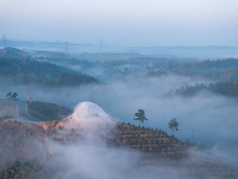 Tower emerging from low cloud: the white membrane form visible above the mist, terraced slopes below