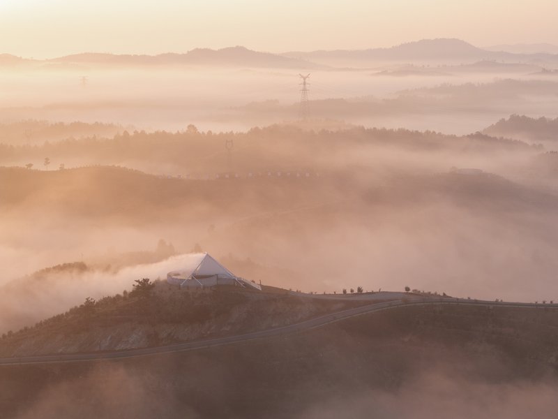 Dawn aerial: the membrane-wrapped tower rising from morning fog over the terraced tea-oil hillside