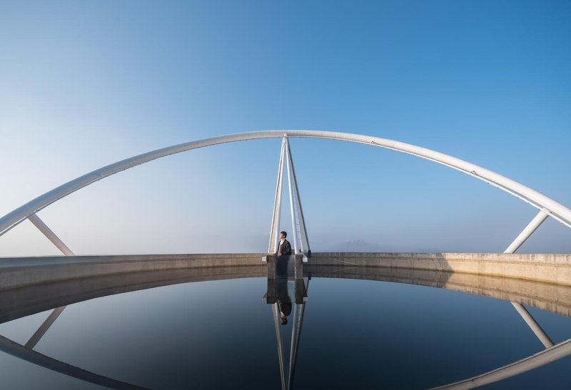 Walkway on the reservoir: figure walking across the mirrored staircase toward the water, steel arch overhead