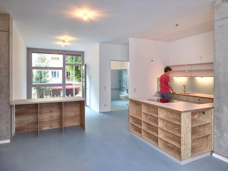 Open kitchen and living area with plywood cabinetry, grey flooring, and exposed concrete columns