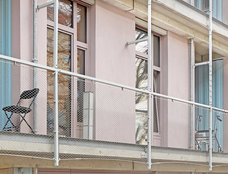 Narrow balcony with white mesh railing, folding chair, and pale pink facade with teal window frames