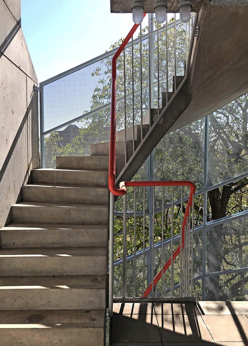 Concrete staircase with red metal handrail and perforated metal balustrade backed by green foliage
