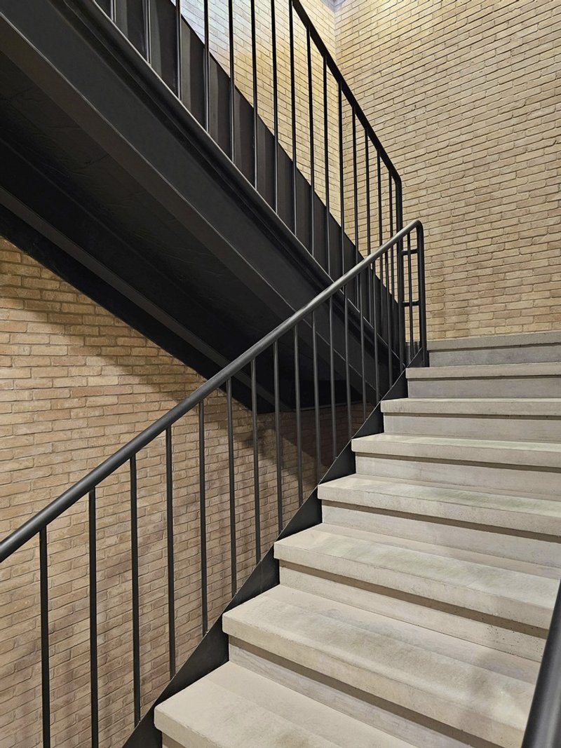 Stone staircase with black steel balustrade ascending beside a textured brick wall in afternoon light