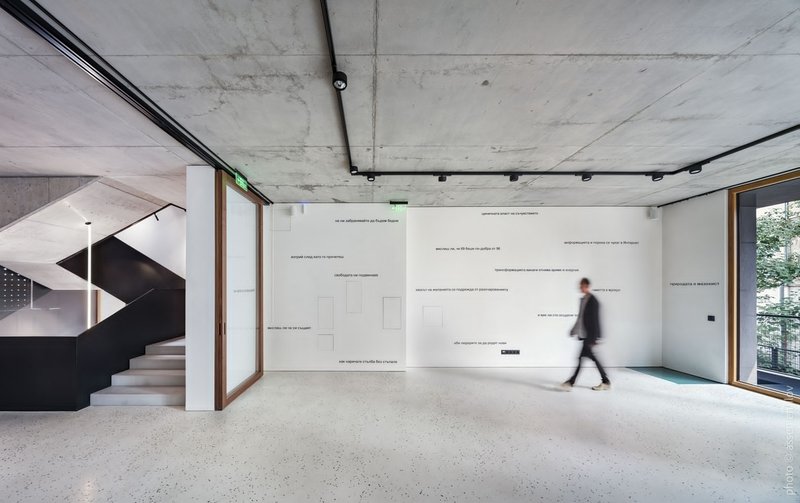 Open gallery space with polished concrete floor, text-filled wall display, and person walking past glazed opening