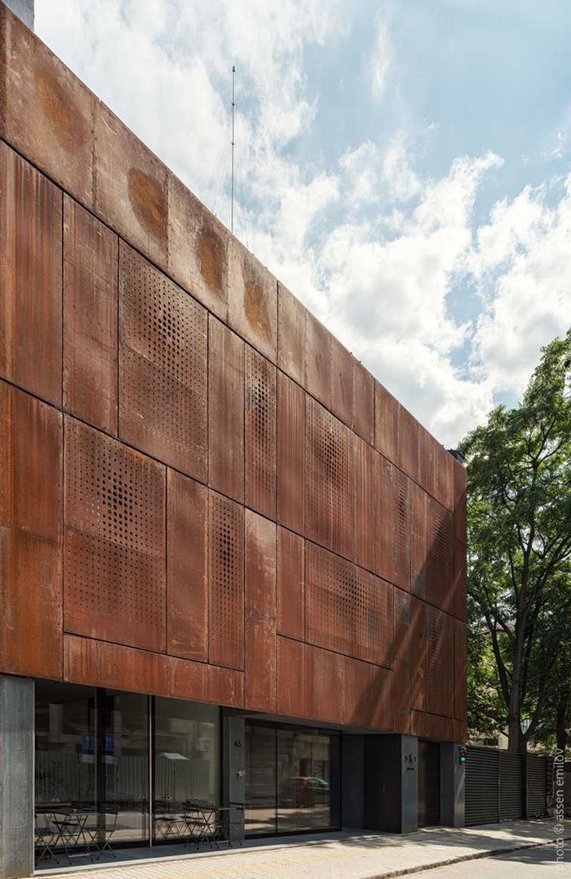 Weathered corten steel facade with perforated panels above glazed ground floor entrance and adjacent tree canopy