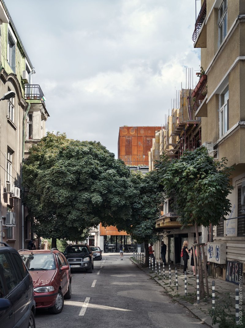 Distant view of the corten steel volume framed by street trees and neighboring residential buildings