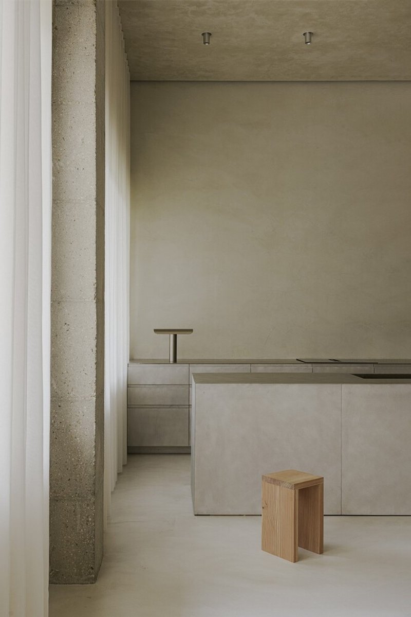 View through concrete columns toward a pale plastered kitchen counter with a timber stool on polished flooring