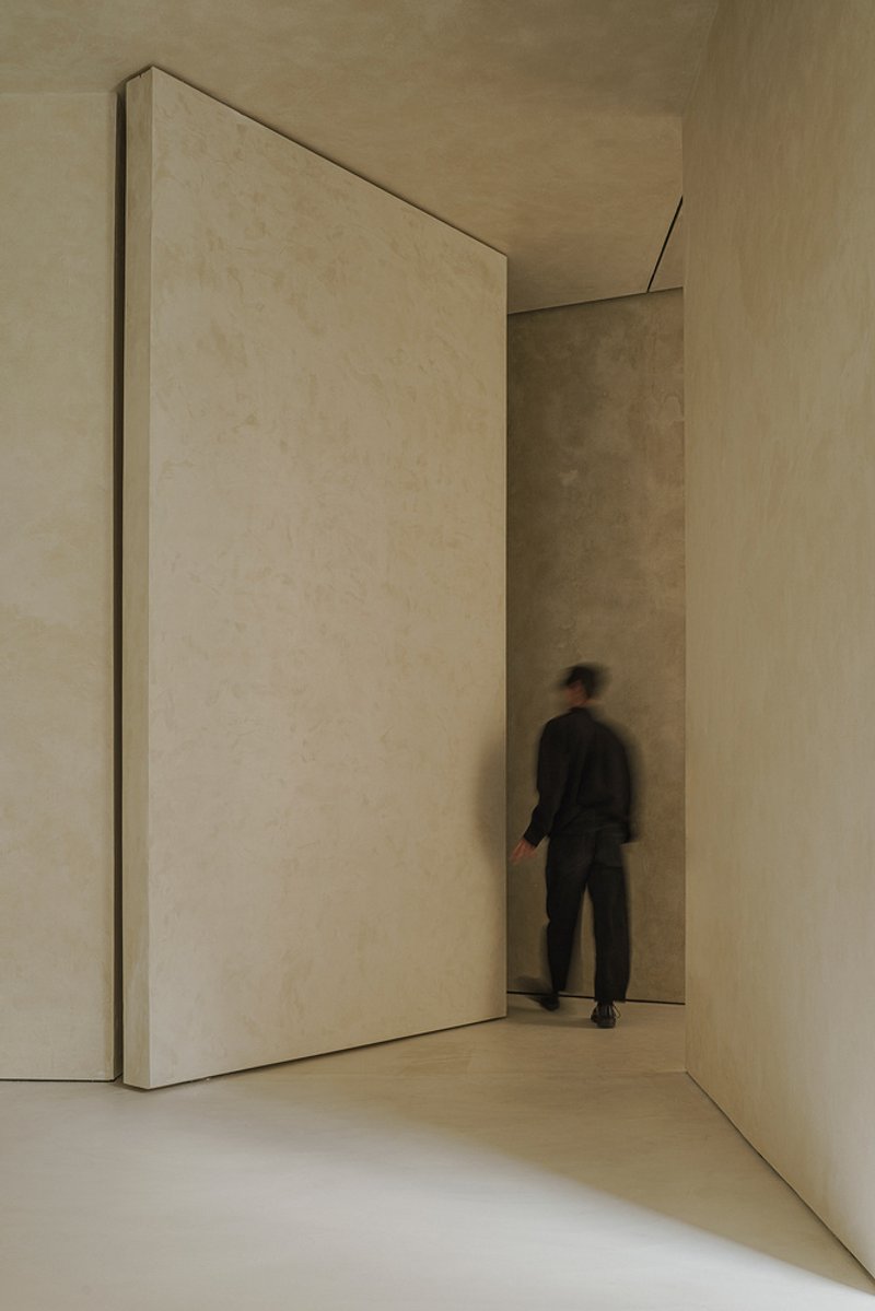 Tall pivoting plaster-clad door with a person walking through the narrow passage in soft light