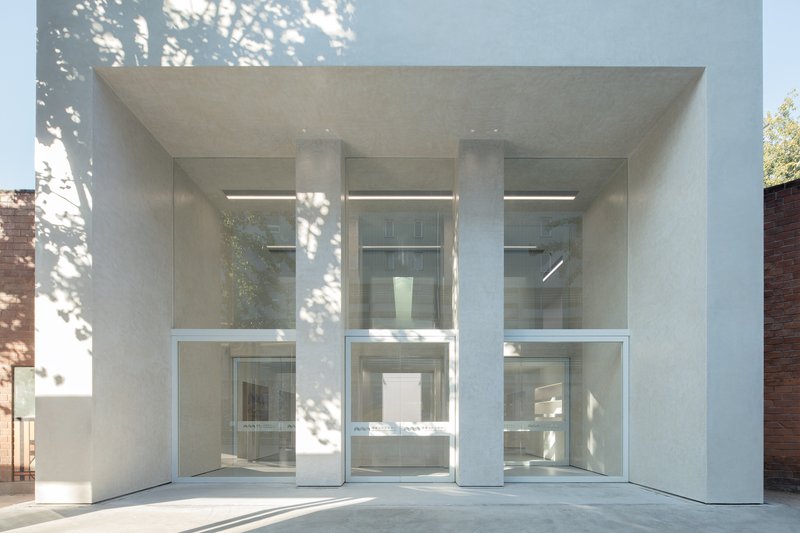 Entry close-up: three full-height glass panels between white concrete columns, tree shadows on the facade, double-height lobby visible