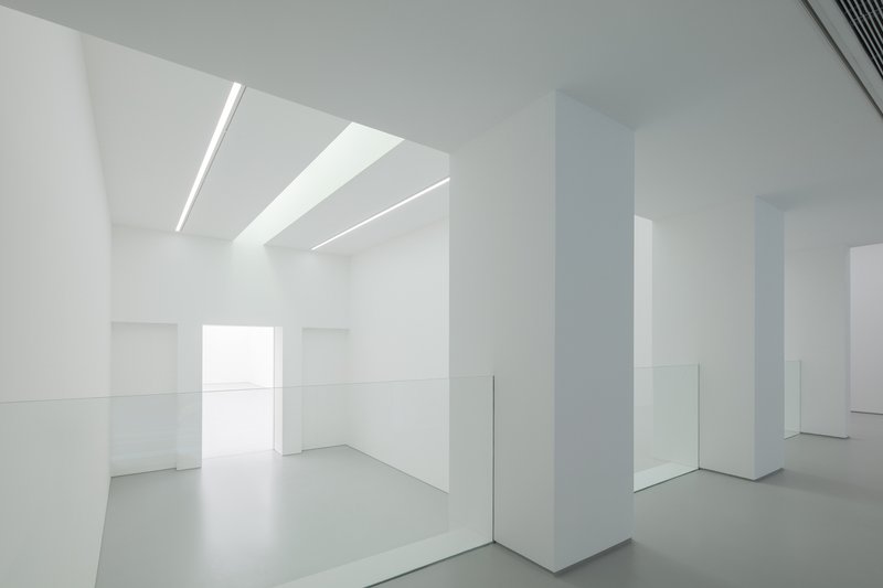 Upper level interior: white columns, glass balustrade, linear ceiling light, volume open to the floor below, window at the far end