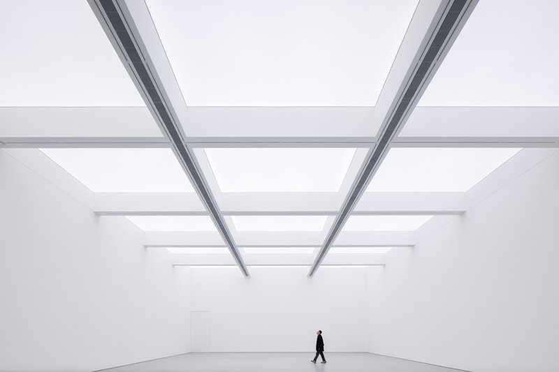 Main hall with figure: person walking across the vast white room under a skylight grid, steel beams converging above
