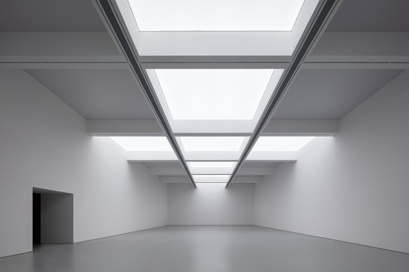 Main exhibition hall ceiling: full-width skylight grid, steel beams, white walls, dark doorway at left