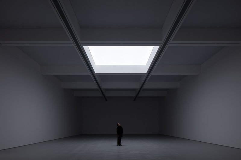 Darkened hall: single central skylight illuminating a dark exhibition space, figure standing below