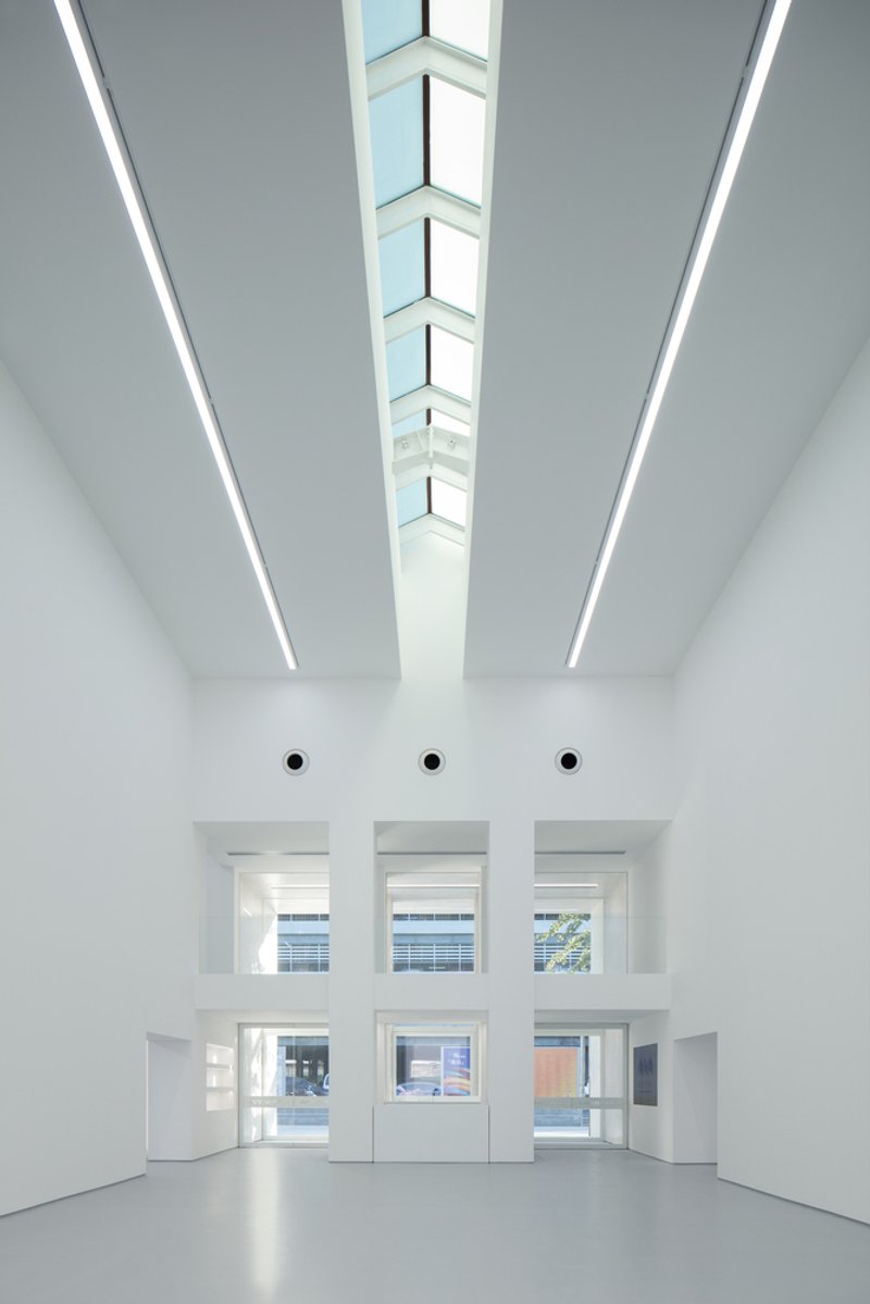 Lobby interior: tall white volume, skylight strip running the length of the ceiling, three windows to the street at the far end