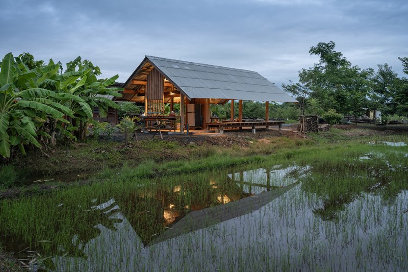 Open timber pavilion with metal roof reflected in flooded rice paddy at dusk