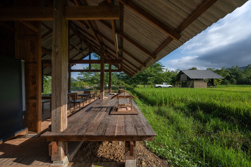 Interior view through exposed timber frame opening onto green rice fields under cloudy sky