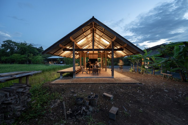 Gabled timber pavilion with corrugated roof illuminated from within at dusk in a rural setting