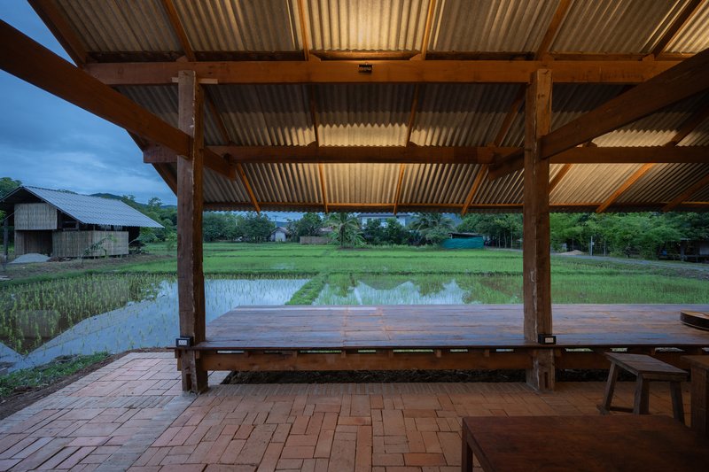 Covered terrace with timber columns, corrugated metal roof and brick paving overlooking rice paddies at dusk