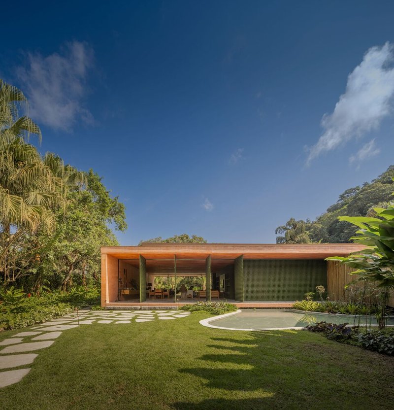 Single-story timber pavilion with large glazing opening onto a lawn and curved paver path under blue sky